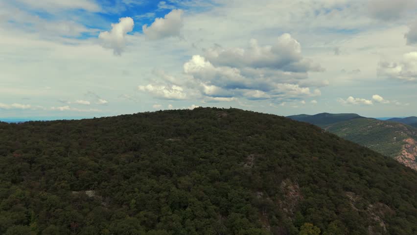 An aerial view over Storm King Mountain, NY on a sunny day with blue skies and white clouds. The drone camera dolly in and boom up over the mountain top to reveal the Hudson River and the horizon.