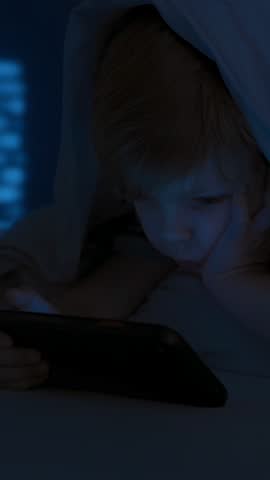 Little boy lying under blanket in dark room, holding smartphone up while staring at screen. Digital addiction, screen dependency, gadget overuse concept