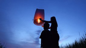A mother and child release a Yi Peng lantern during Thailand’s Loy Krathong festival at night. The glowing lantern rises into the dark sky, symbolizing letting go of bad luck and welcoming good fortun - Powered by Shutterstock - Get 15% off with code: PIKWIZARD15
