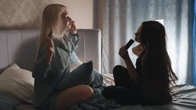 Two siblings enjoy playful bedtime moment on bed as girl in black pajamas sings into remote control like microphone while her sister dances along - Powered by Shutterstock - Get 15% off with code: PIKWIZARD15