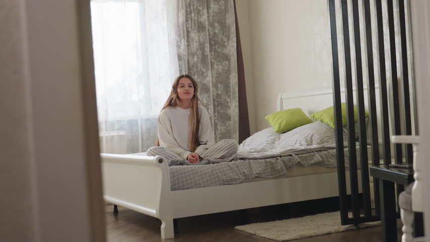 Student seated on bed smiles warmly watching roommate walk into room wearing elegant black dress, admiring her fashion style, bright daylight through window adds soft glow to modern interior