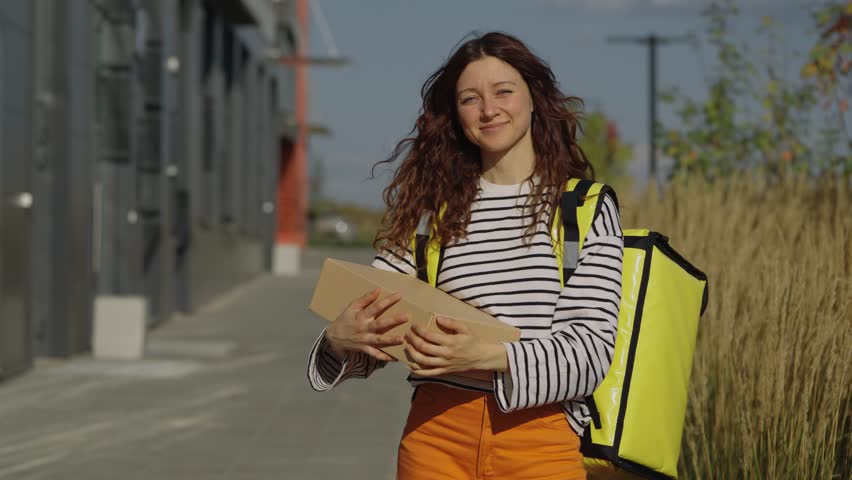 Young female courier smiling and making eye contact while walking through a city street carrying a cardboard package and wearing a thermal delivery backpack, upbeat urban delivery scene
