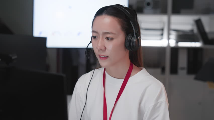 Young Asian call center woman wearing headset and microphone while working at computer in modern office. Professional customer support representative providing online assistance, technical help