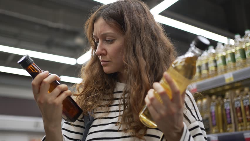 Thoughtful curly-haired woman comparing two different bottles of olive oil in a grocery store, carefully reading the labels and ingredients to make the best choice for a healthy diet