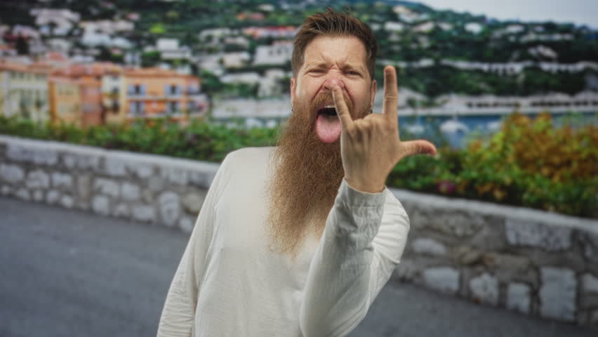 Man with long beard makes rock horns gesture with hand while sticking out tongue on beach promenade; spirited enthusiasm.