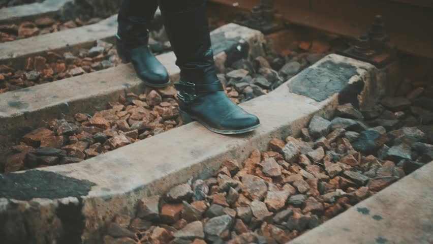 Close-up shot legs walking along railroad track, feet stepping on sleepers, unrecognizable silhouette person in grunge black boots, rocker style with chains, back rear view woman goes on adventure 4k.