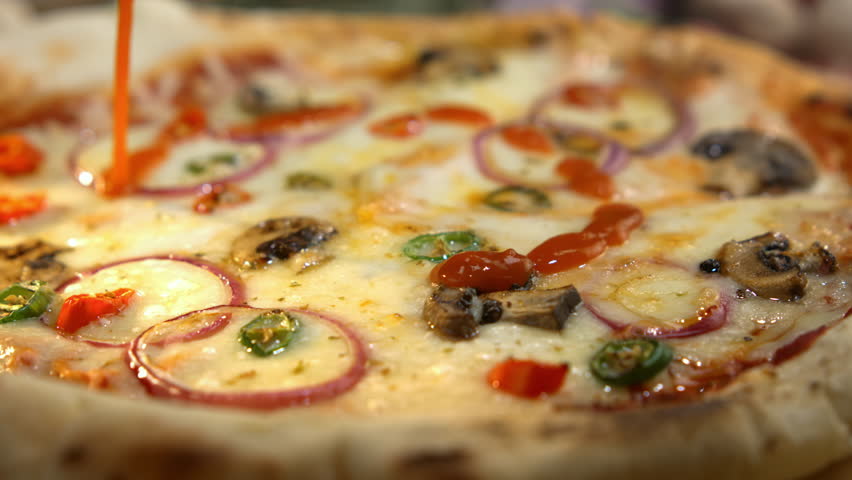 Delicious hot pizza with mozzarella cheese, mushrooms, and spicy peppers receiving a final touch as red tomato ketchup is poured in a spiral pattern before serving in a restaurant