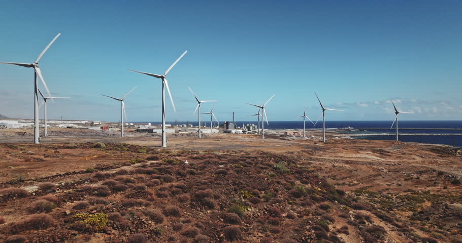 Tenerife: Wind farm turbines generating clean, sustainable power on a coastal landscape, with industrial structures and the blue Atlantic Ocean under a clear sky, Canary Islands. Drone flight footage