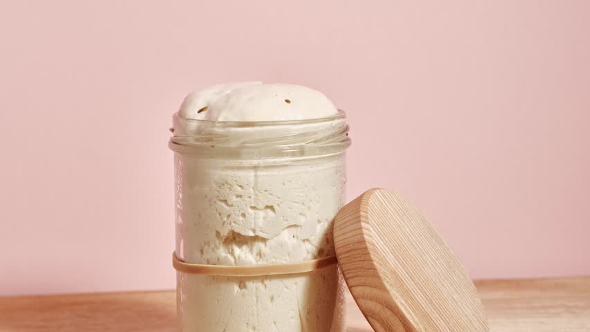 Wheat sourdough levito madre rising in time-lapse with a domed top forming above the rim of the glass jar as fermentation pushes the dough upward, wooden lid placed aside