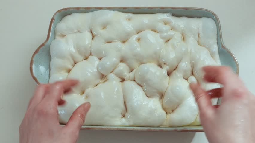 Close-up of raw focaccia dough in baking tray being massaged with hands, glistening with olive oil and forming air bubbles