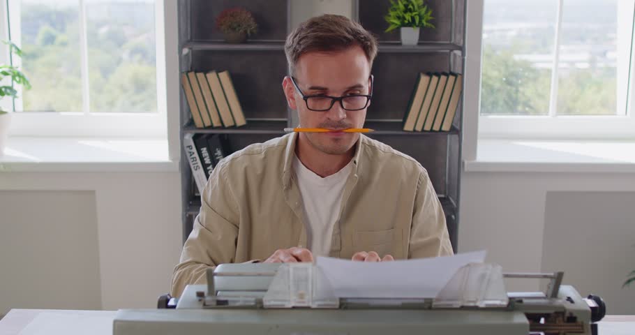 Busy writer typing on vintage typewriter in office by windows, holding pencil in mouth. Man wearing glasses for focus and creativity of retro vibes, professional authors routine working on old machine