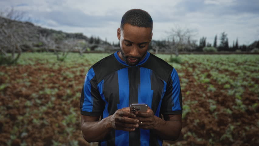Man checking smartphone with fist pump gesture and smiling while standing in a countryside field; joy victory.