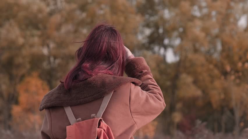 Teen girl standing with her back to the camera, turning around with a gentle smile, wearing sunglasses and a warm brown coat in an autumn landscape.