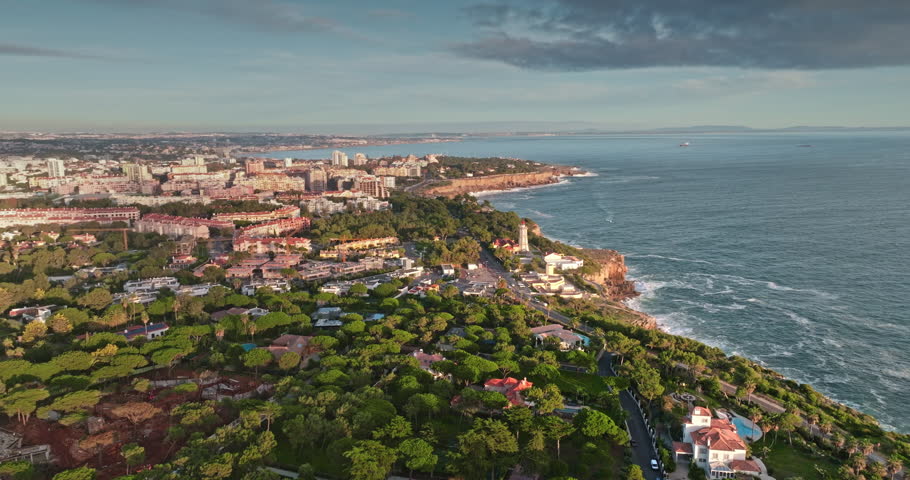 Portugal, Lisbon: Cascais city skyline extends along rugged Atlantic Ocean coastline, residential buildings, lush green trees, and the Santa Marta lighthouse at sunset. Aerial view drone panorama