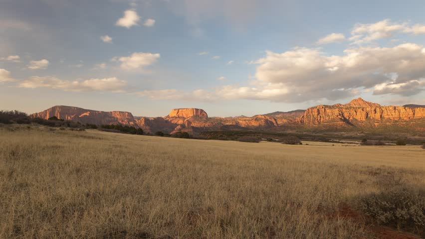 4k time lapse video of clouds drifting gently from left to right above an open grassy field framed by the distant cliffs of Kolob Canyon, Zion NP Utah, USA as the warm evening sun fades to dusk.