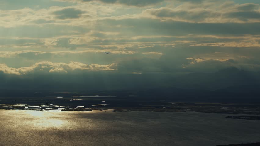 View of passenger plane in cloudy sky at sunset and silvery surface of sea bay with mountain ranges on shore and backwaters in valley. Slow motion