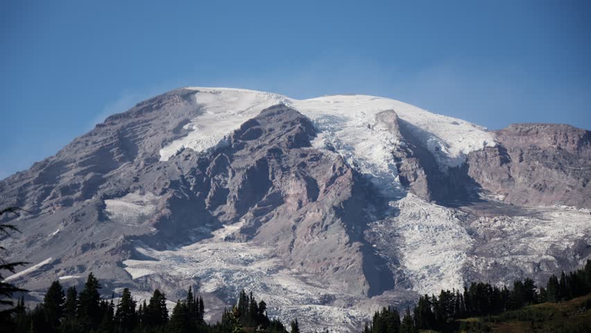 Snow-covered mountain peak with a clear blue sky above and silhouettes of trees below. Beauty in nature.