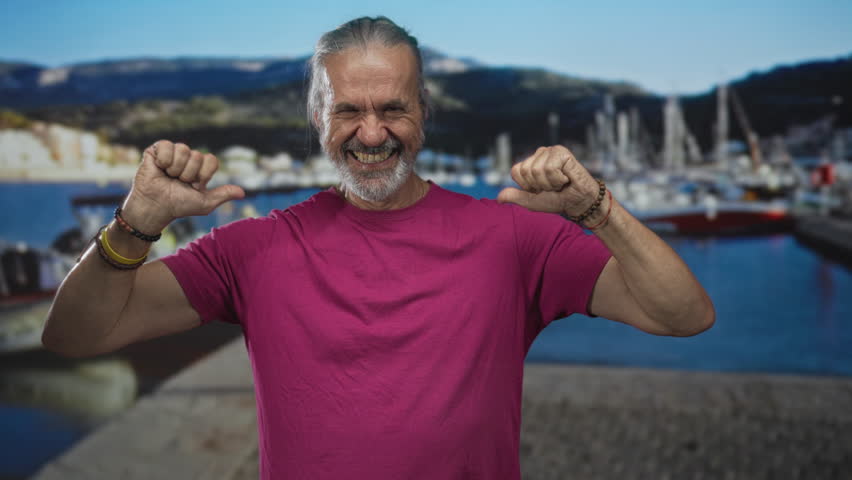 Man with thumbs pointing to chest and flexing arms on street by a harbor pier with moored boats, smiling toward camera; self confidence.