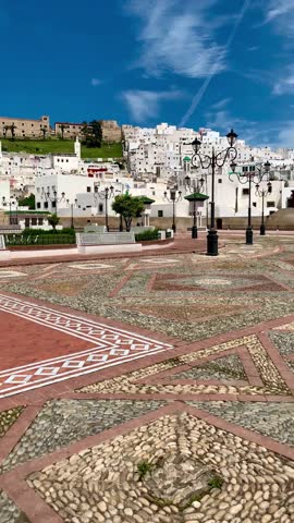 Tetouan, Morocco: Ornate white pavilion with arched design and green pyramidal roof stands in Feddan Park square amid intricate red mosaic tile flooring, palm trees, and ornate lamps