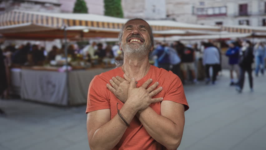 Middle aged man with hands crossed over chest and pointing fingers toward camera in street market; joyful gratitude.