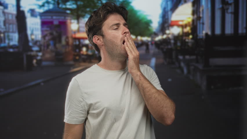 Man, young, wearing white t shirt, touching chin with right hand and bare forearm visible, standing on a busy city street at dusk with blurred cafes; thoughtful concern.