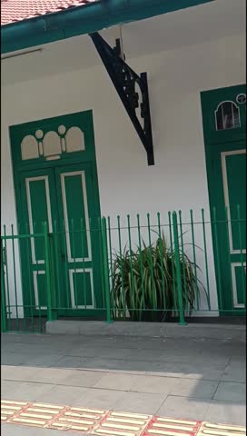 Building with white walls and green doors and windows, featuring a matching green metal fence in front. The architectural design has a vintage or colonial style.