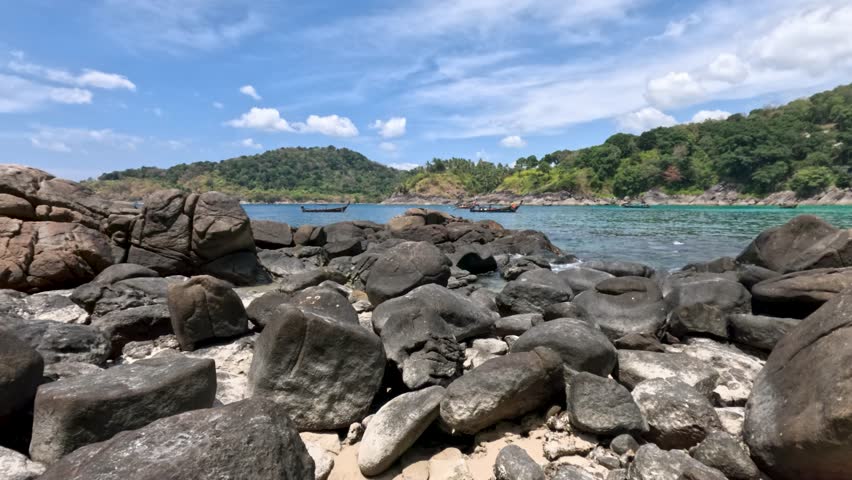 Camera slowly pans rocky beach with turquoise water, green hills, bright daylight, and blue sky