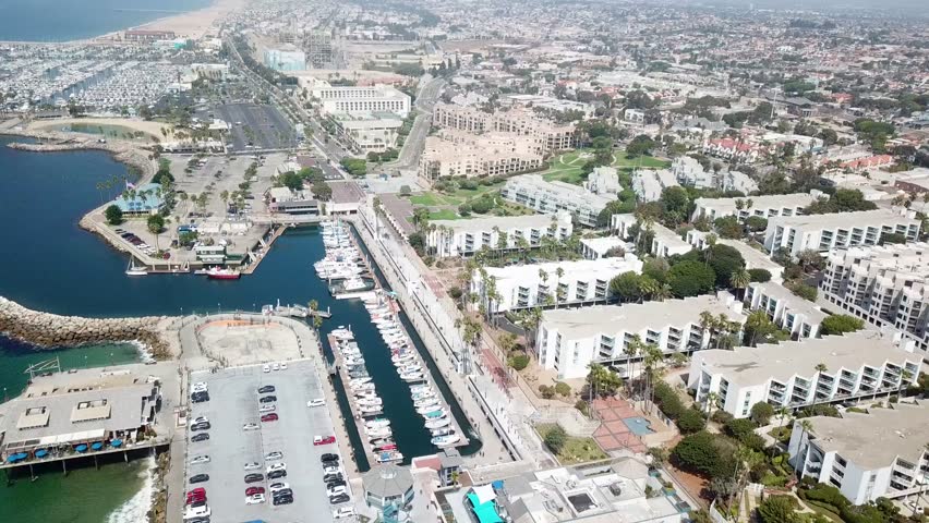 Drone pulls back and pans over the harbor channel at Redondo Beach with docked boats waterfront apartments and parking areas leading toward the inland city. Clear coastal light and strong geometry.