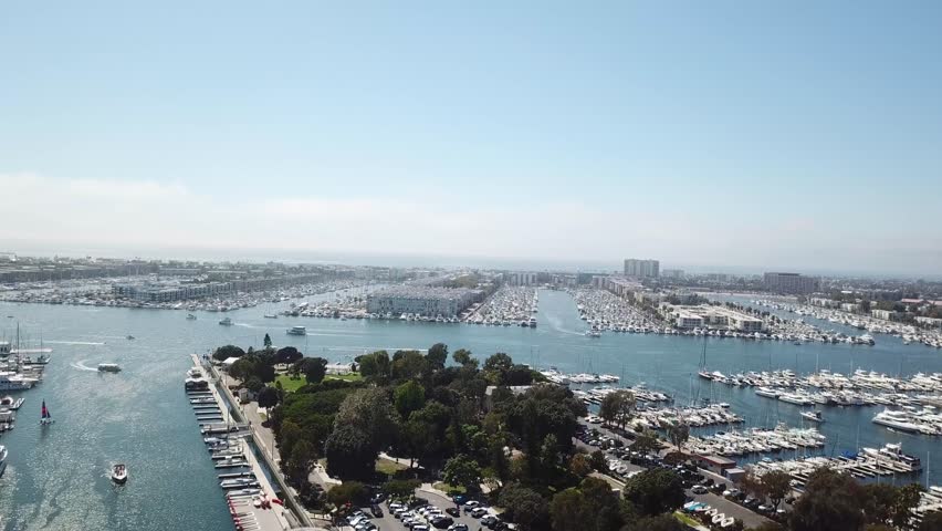 Wide aerial of Marina del Rey, CA. Camera pushes in and ascends, revealing long marina basins packed with yachts, a central navigation channel, and waterfront development under clear SoCal skies.