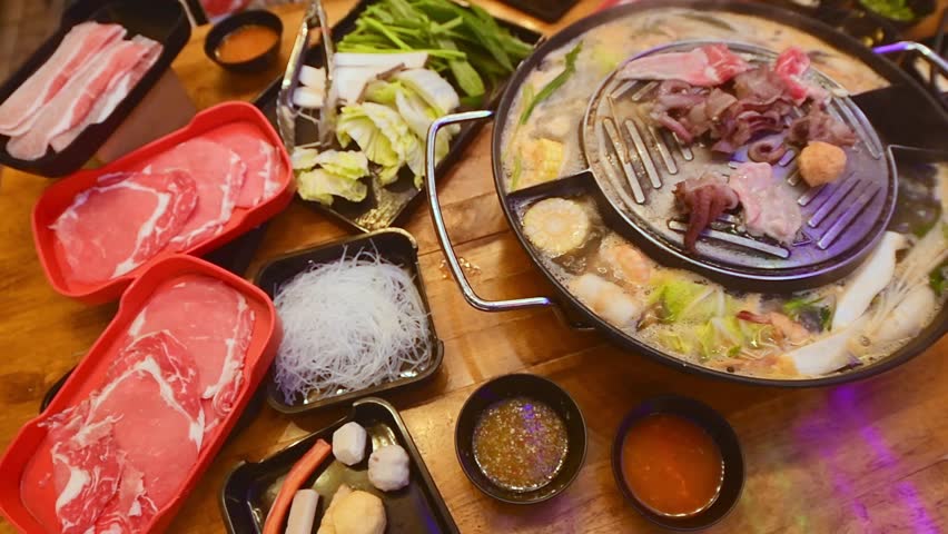 Top view of traditional Thai BBQ hotpot (Moo Krata) with mixed seafood, octopus, shrimp, fish balls, noodles, mushrooms, cabbage, corn, garlic, and dipping sauces on wooden table in Bangkok, Thailand.