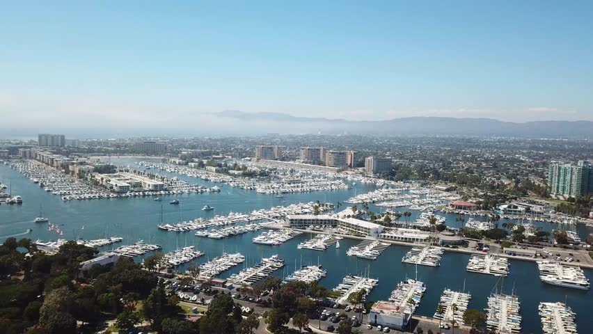 Aerial slow push-in with a pan across Marina del Rey, Los Angeles. Reveals dense marina basins filled with yachts, waterfront condos and hotels, and the Westside skyline under clear blue skies