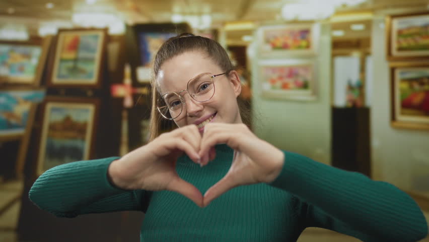 Woman forming heart shape with hands in colorful art gallery building surrounded by paintings; affection.