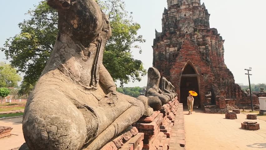 Ayutthaya, Thailand – March 2023: Woman in traditional Thai dress holding yellow umbrella walking by headless Buddha statues at Wat Chaiwatthanaram, UNESCO World Heritage Site symbol of ancient Siam.