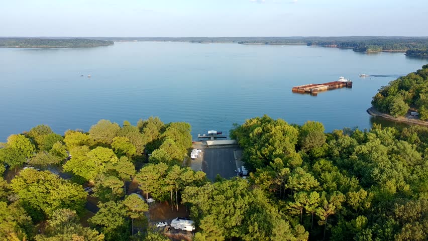 A beautiful lake with a boat in the water. The boat is a large one and is in the middle of the lake. The lake is surrounded by trees and there are several cars parked near the shore