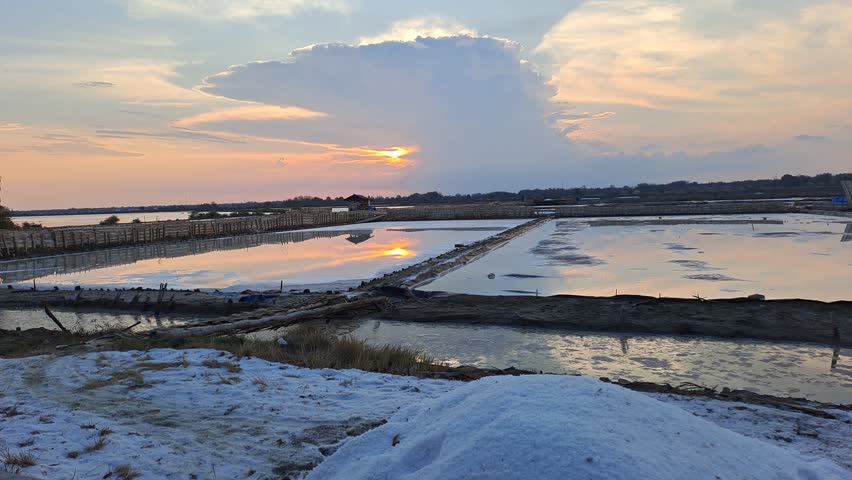 Peaceful sunset over a traditional salt farm, glowing in warm orange and red hues, with calm reflections and beautiful clouds painting the serene evening sky.