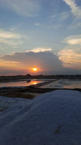 Peaceful sunset over a traditional salt farm, glowing in warm orange and red hues, with calm reflections and beautiful clouds painting the serene evening sky.