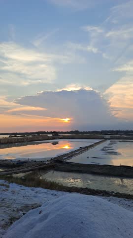 Peaceful sunset over a traditional salt farm, glowing in warm orange and red hues, with calm reflections and beautiful clouds painting the serene evening sky.