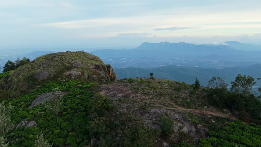 Scenic mountain peak view with people enjoying the panoramic landscape at sunrise. Lush green valleys, distant hills, and a peaceful lake under a serene sky. Feels like the top of the world.