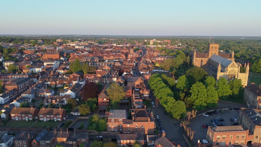 A cinematic parallax drone shot captures the picturesque cityscape of St Albans bathed in warm afternoon sunlight, highlighting historic architecture and charming streets.