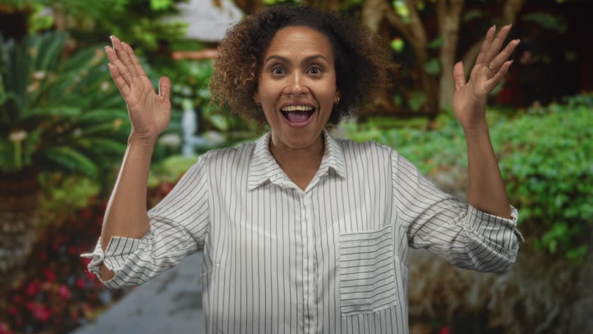 Woman smiling with hands raised and open palms in a forest setting among greenery and park foliage, wearing a striped shirt; joy celebration.