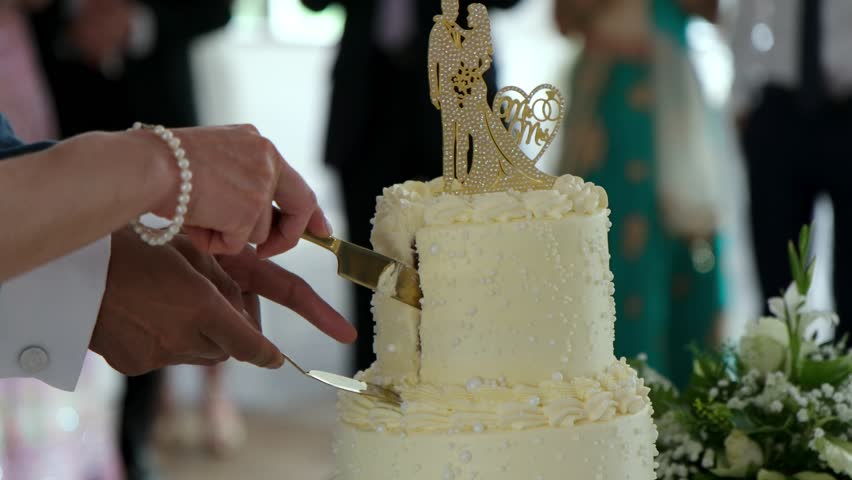 Elegant newlywed couple slicing white tiered wedding cake with gold bride-groom topper. Bride wears pearl bracelet. Perfect for romantic celebrations, receptions, or matrimony content.