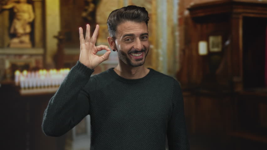 Young smiling man shows ok sign with hand in candlelit ancient building corridor indoors; approval.