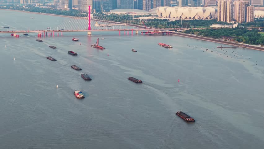 Cargo ships and boats navigate the Qiantang River in Hangzhou China. A red bridge spans the river, with the city skyline visible in the distance