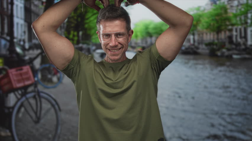 Man smiling and raising both bare forearms beside canal bicycles on amsterdam street during daylight, wearing glasses and green tshirt; joy travel.