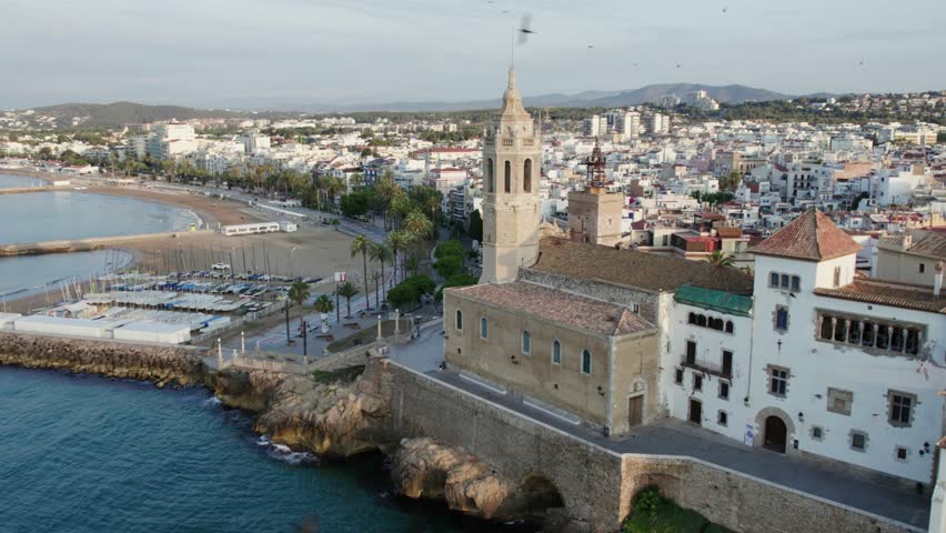 Aerial view of the Church of Sant Bartomeu and Santa Tecla, with its bell tower, standing tall by the coast, contrasted against the cityscape, Sitges, Barcelona, Spain.