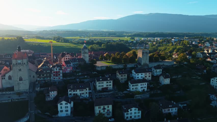 Aerial view of Orbe cityscape with towers and church, bathed in the warm glow of sunset, amidst rolling hills and lush greenery, Orbe, Vaud, Switzerland.