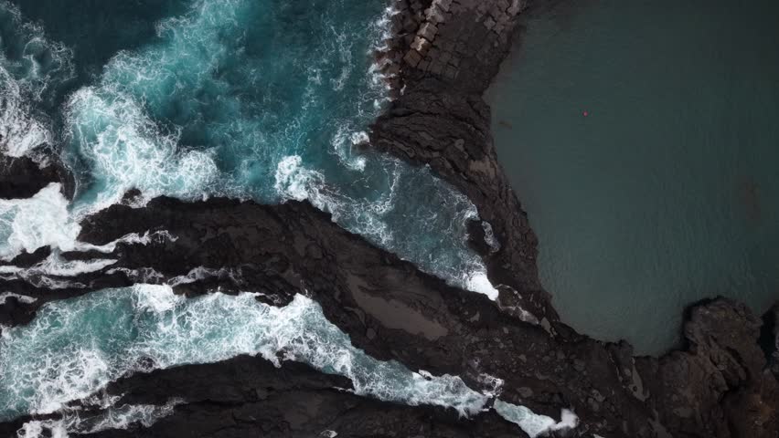 Aerial view of a dramatic coastline where turquoise waves crash against dark, jagged rocks, creating a mesmerizing contrast of color and texture, Seixal, Portugal.
