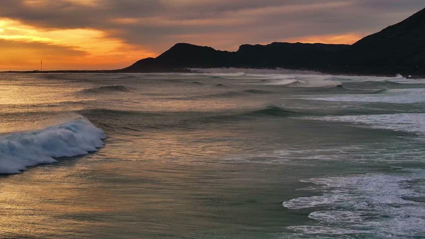 Aerial view of powerful waves crashing onto the shore at sunset, as the golden light touches the dark mountain silhouette, Cape Town, Western Cape, South Africa.