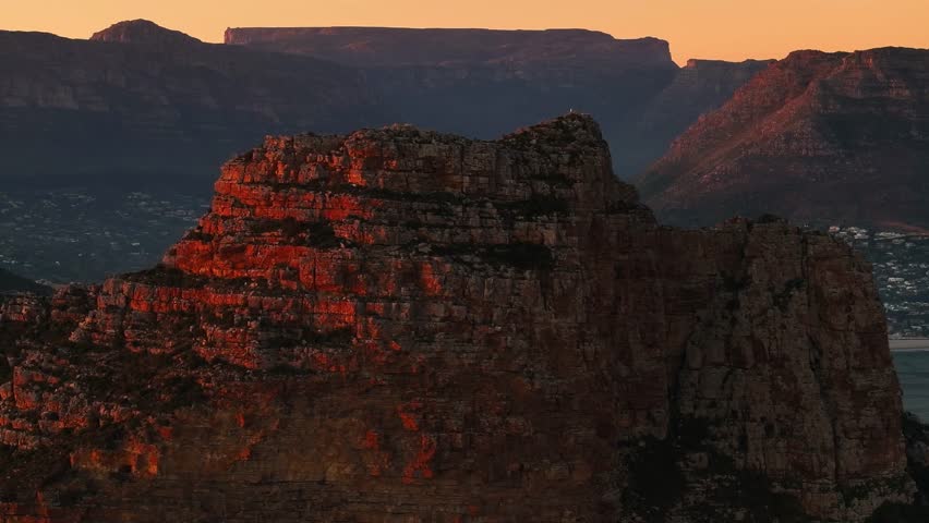 Aerial view of a rugged mountain range bathed in the warm glow of sunset, with Table Mountain visible in the background, Cape Town, Western Cape, South Africa.