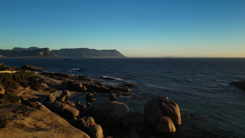 Aerial view of rugged coastline boulders, reflecting the golden light of the setting sun against the deep blue ocean, Cape Town, Western Cape, South Africa.